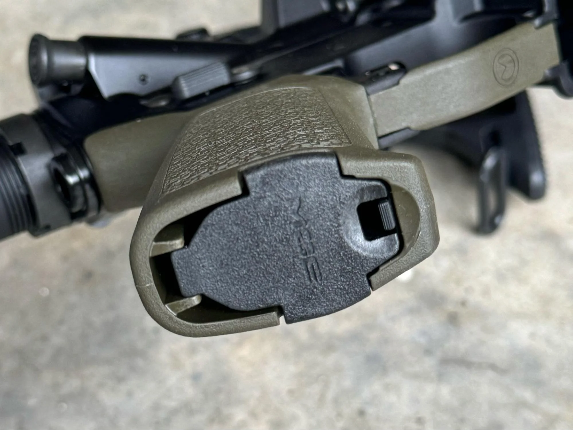 A close-up photo focusing on the grip latch of an AR-15 pistol grip, highlighting the gap around the latch, suggesting a design improvement