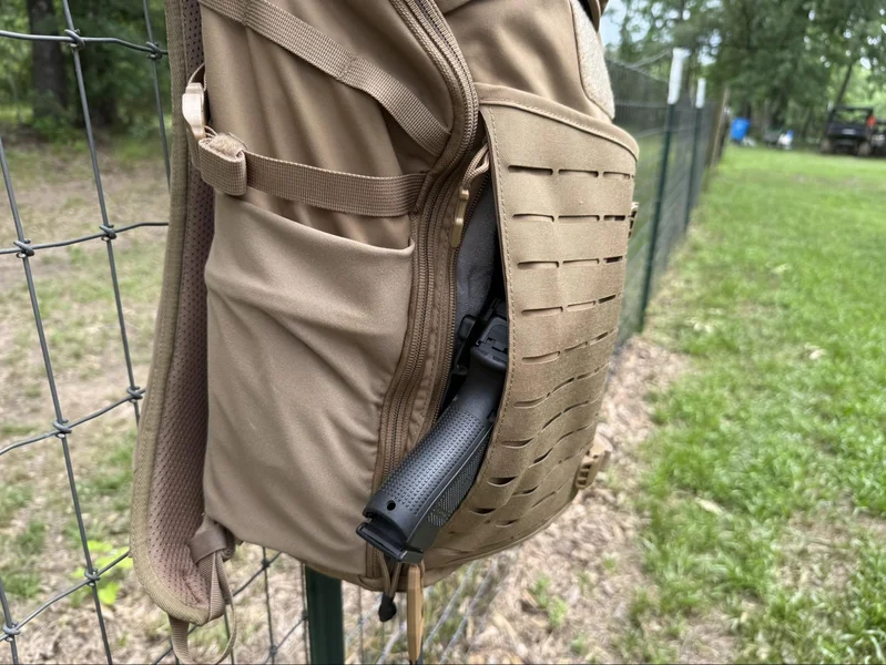 Close-up of Vertx Siege 25L backpack’s concealed carry pocket partially unzipped, showing a holstered handgun for demonstration purposes.