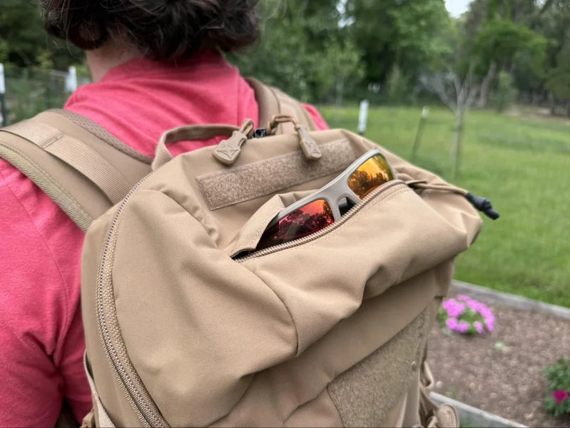Close-up of sunglasses stored in the dedicated top pocket of the Vertx Siege 25L backpack, worn by a person in a red shirt