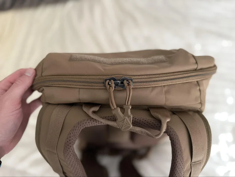 Close-up of the top of the Vertx Siege 25L backpack showing YKK locking zippers and padded shoulder straps, with a hand holding the bag.