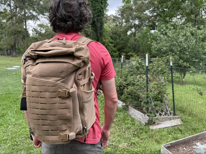 Man wearing a tan Vertx Siege 25L backpack in a backyard garden, showing the bag’s tactical build and size from behind.