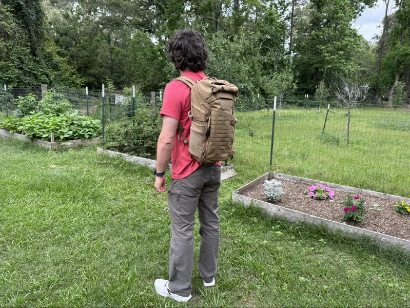 Man wearing a tan Vertx Siege 25L backpack standing in a backyard garden, facing away from the camera, demonstrating the pack’s size and fit for daily carry.