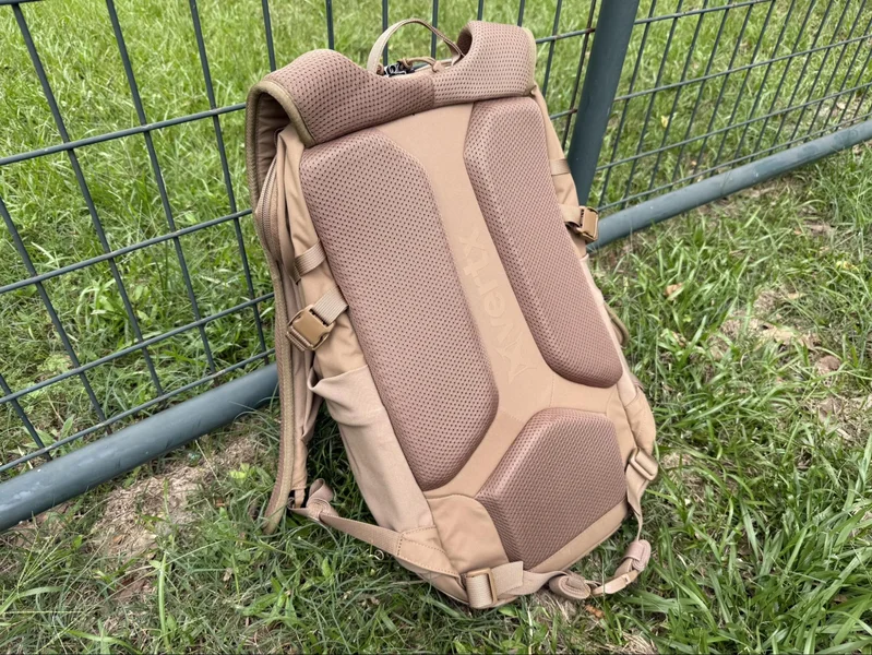 Rear view of the Vertx Siege 25L backpack showing padded back panel and shoulder straps, resting against a metal fence on grass.