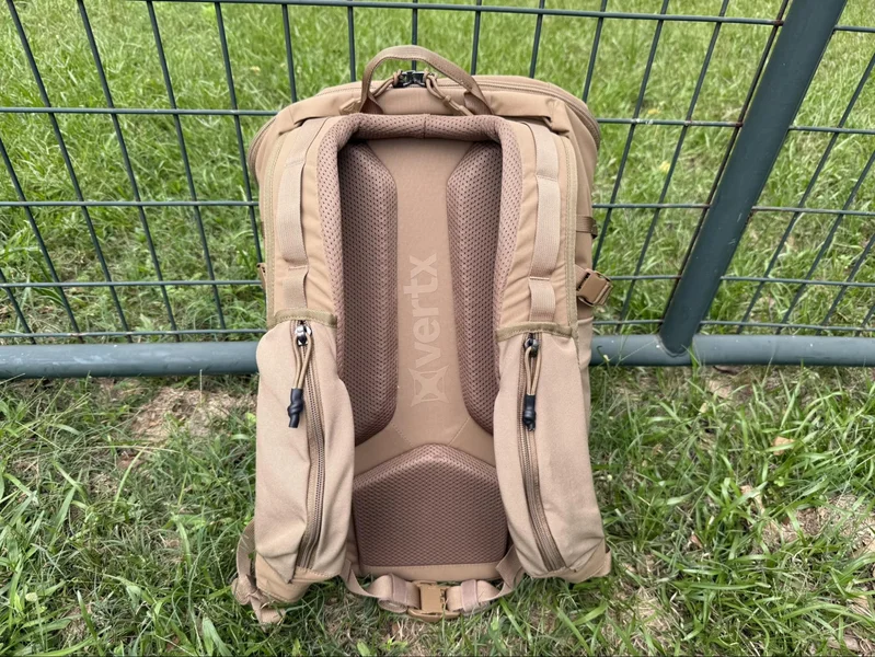 Rear view of the Vertx Siege 25L backpack showing padded shoulder straps, sternum strap, and ventilated back panel resting against a fence.