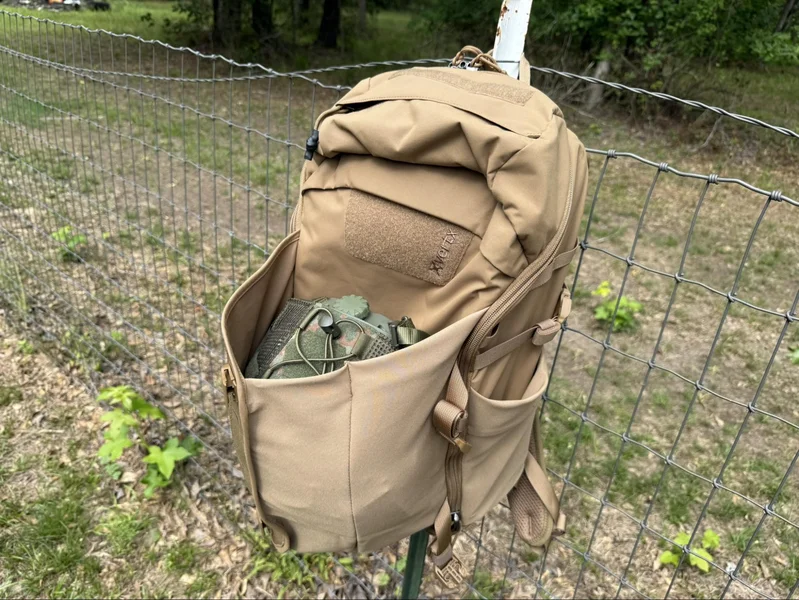 Vertx Siege 25L backpack hanging on a fence with expandable front pocket open, holding gear and showcasing storage capacity for items like a helmet or lunch.