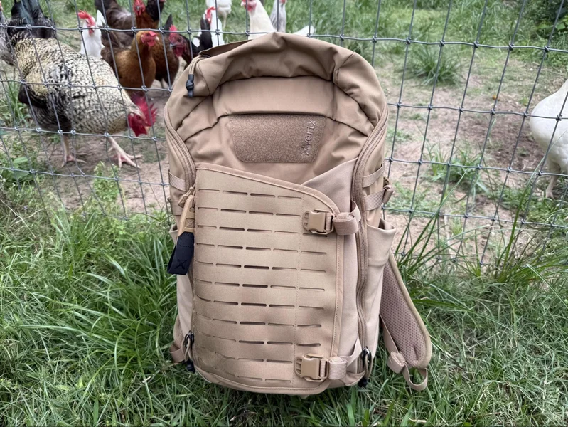 Vertx Siege 25L backpack standing upright in grass against a wire fence, with chickens in the background highlighting its rural, everyday carry appeal.