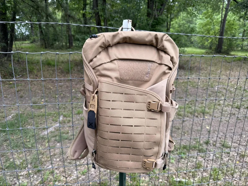 vertx siege 25l Tan tactical backpack hanging on a wire fence outdoors, designed for everyday carry with MOLLE-style webbing and buckled straps