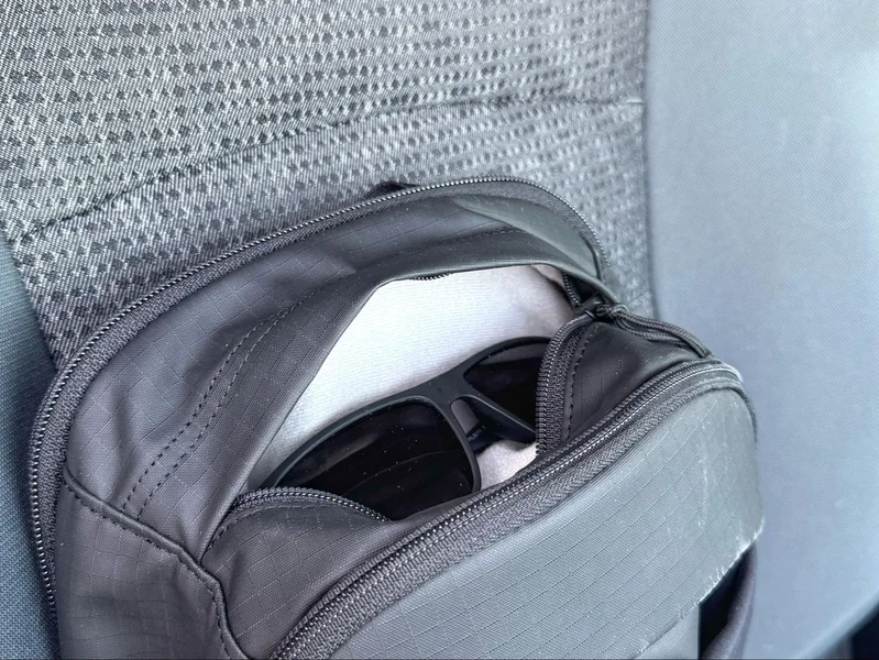 Top pocket of sling bag holding a pair of black sunglasses, shown inside a car