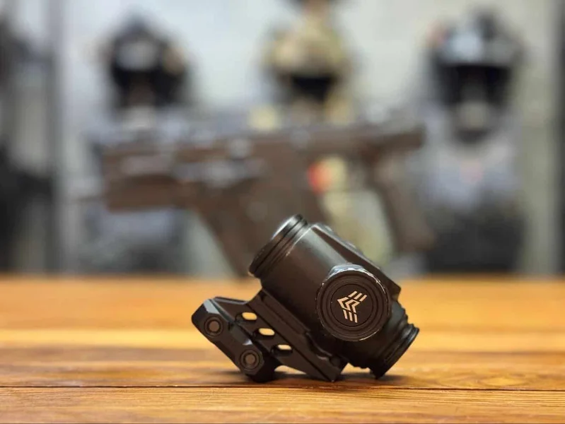 Close-up of Swampfox Raider 1X prism optic resting on a wood surface, tactical helmets blurred in the background.