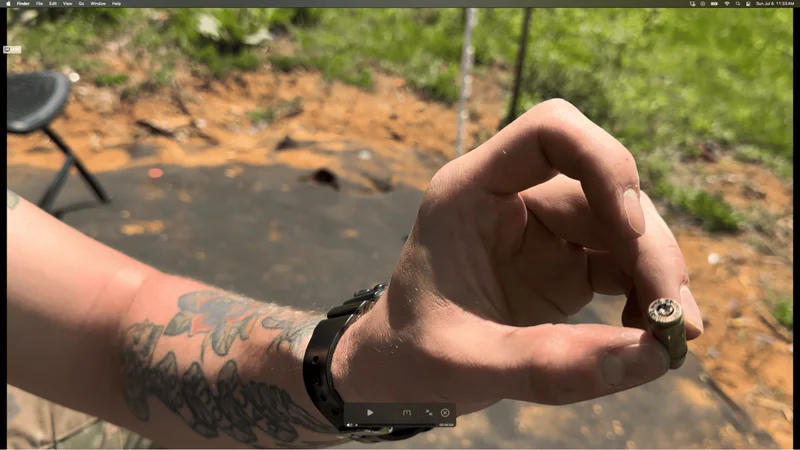 Close-up of a hand holding a ruptured bullet casing at an outdoor shooting range, with dirt and grass in the background.