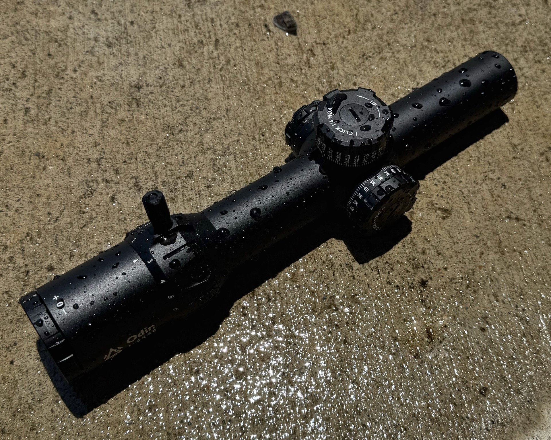 GUNNR Odin LPVO rifle optic lying on wet concrete, covered in water droplets after a simulated rain test