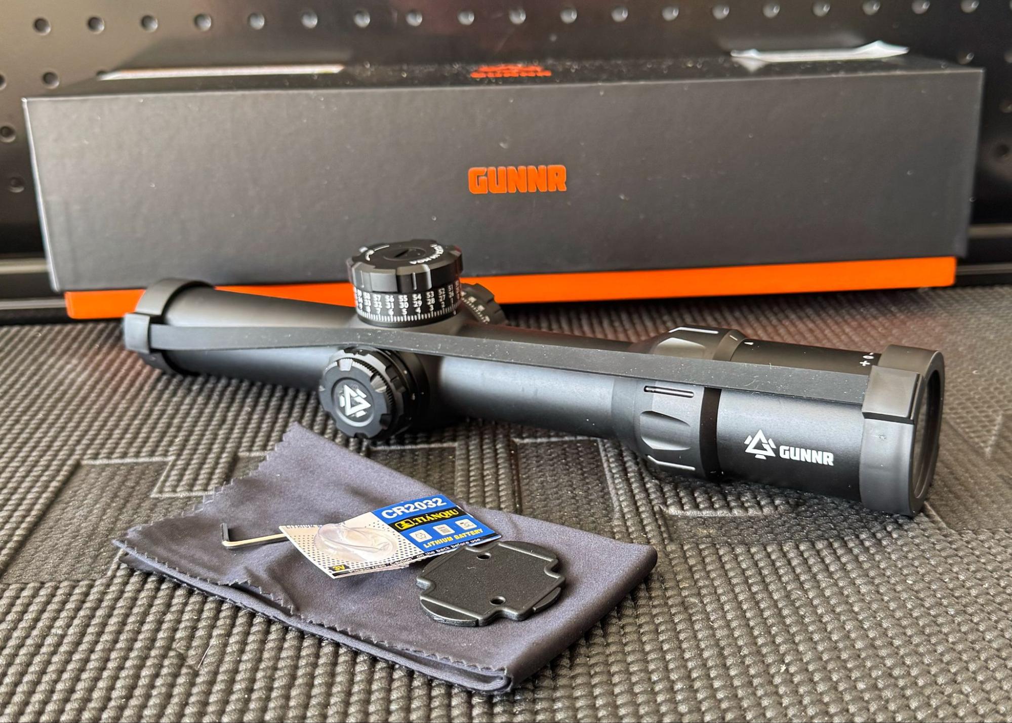 GUNNR Odin LPVO rifle optic with included accessories on a table, including throw lever, torque key, CR2032 battery, turret cap tool, lens covers, and lens cloth, with branded box in background