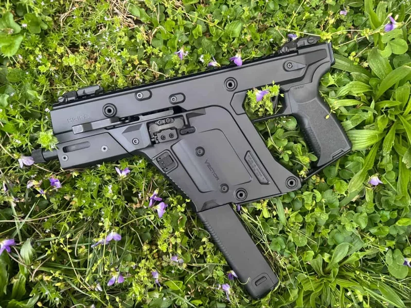 Kriss Vector Gen 3 9mm pistol resting on grass with purple flowers, showing side profile of firearm including trigger, magazine, and rail attachments.