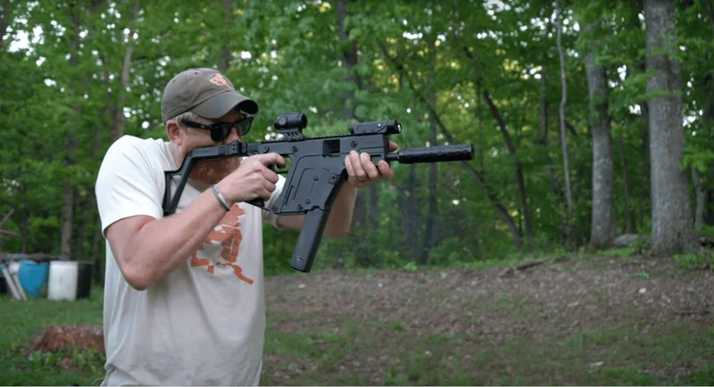 Man aiming a Kriss Vector Gen 3 9mm rifle with suppressor and optic in an outdoor wooded area.