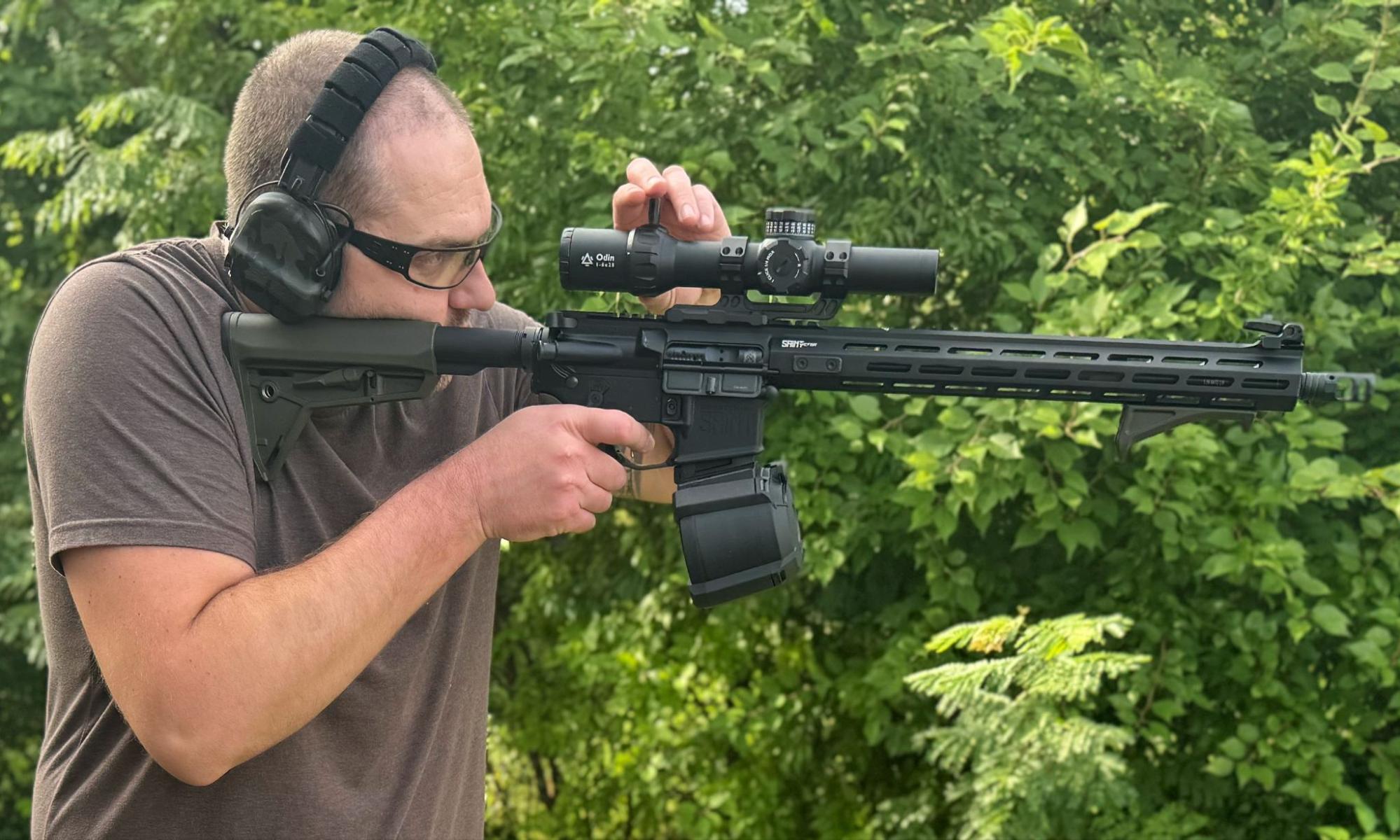 Man aiming a rifle equipped with the GUNNR Odin LPVO, featuring an installed throw lever, wearing hearing protection and shooting glasses outdoors