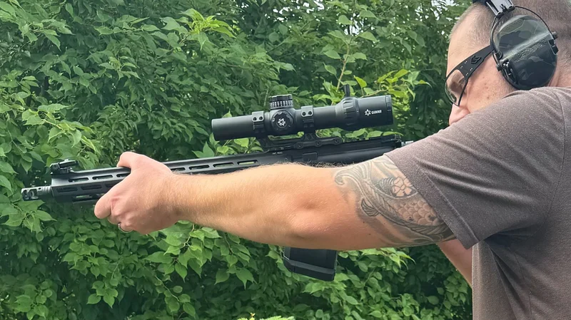 Man aiming a rifle outdoors, equipped with a GUNNR Odin 1-6x28 LPVO optic, wearing hearing protection and sunglasses