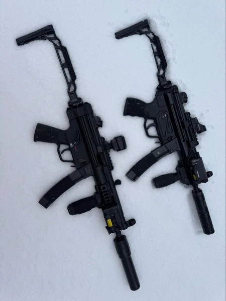 Two Military Arms Corp MAC5K subguns with suppressors and optics lying side by side on snow.
