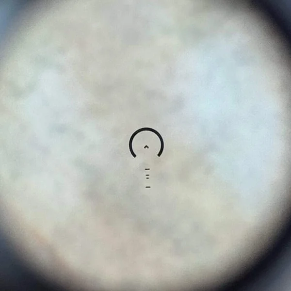 View through the GLx 1x MicroPrism optic showing illuminated chevron reticle with auto-ranging ladder on a blurred background.
